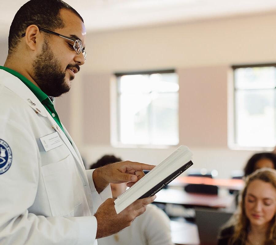 a male student standing in a classroom reading from a textbook